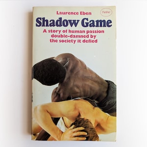 Puede incluir: Una portada de libro con una foto de dos hombres luchando. El título del libro es "Shadow Game" y el subtítulo es "Una historia de pasión humana doblemente condenada por la sociedad que desafió".
