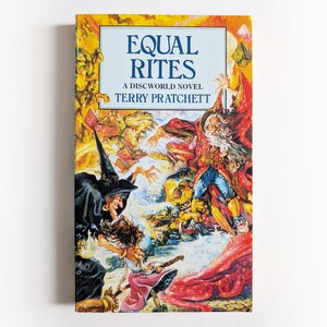 May include: A paperback book titled Equal Rites, A Discworld Novel by Terry Pratchett. The cover features a vibrant, detailed illustration of a wizard in a red robe and a witch in a black hat and cloak, surrounded by swirling magical energy and creatures.