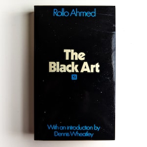 Puede incluir: Un libro negro con letras doradas titulado "The Black Art" de Rollo Ahmed. El libro tiene un logotipo azul y blanco en la portada y una introducción de Dennis Wheatley.