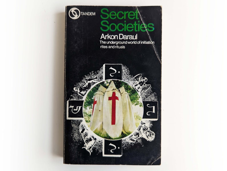 May include: A black and white book cover with the title "Secret Societies" in green letters. The subtitle is "Arkon Daraul The underground world of initiation rites and rituals". The cover features a black and white illustration of three figures wearing white robes with red crosses.