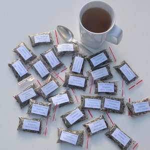 May include: A white mug filled with tea sits next to a spoon and several small plastic bags filled with dried herbs. Each bag has a label with text. The items are arranged on a white surface.