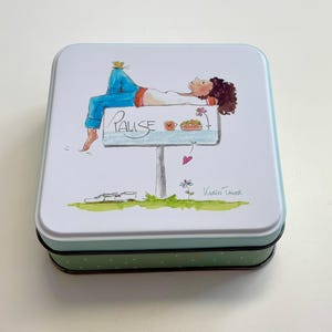 Könnte beinhalten: Eine quadratische Metallbox mit hellblauem Rand und grünem Tupfenboden. Der Deckel zeigt eine skurrile Illustration einer Person, die sich auf einem Schild mit der Aufschrift "Pause" entspannt. Das Kunstwerk umfasst Blumen, ein Herz und die Signatur des Künstlers.