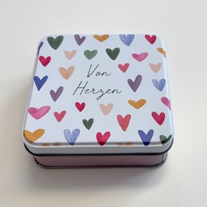 Könnte beinhalten: Eine quadratische Blechdose mit weißem Deckel, verziert mit Aquarellherzen in verschiedenen Farben. Der Text "Von Herzen" ist in schwarzer Schrift geschrieben. Die Dose hat einen rosa Boden.
