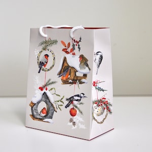 Könnte beinhalten: Eine hellgraue Geschenktüte mit weißen Kordelgriffen, verziert mit einer winterlichen Illustration verschiedener Vögel, darunter Rotkehlchen und Spechte, die auf schneebedeckten Vogelhäuschen, Tannenzweigen und roten Beeren sitzen.