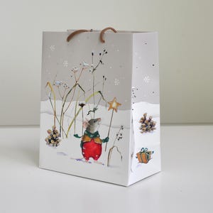 Könnte beinhalten: Eine hellgraue Geschenktüte mit einer Aquarellillustration einer Maus, die einen grünen Schal und eine rote Hose trägt und einen gelben Sternenstab in einer verschneiten Landschaft mit hohen Gräsern, Tannenzapfen und einem kleinen Geschenk hält.
