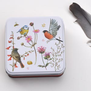 Könnte beinhalten: Eine quadratische, weiße Dose mit Deckel, verziert mit einer Aquarellillustration von Vögeln, Blumen und einer Biene. Die Vögel sind in Braun-, Blau- und Rottönen gehalten. Eine graue Feder liegt daneben.