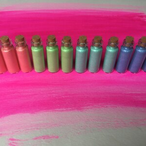 Pode incluir: Uma fila de 14 pequenos frascos de vidro com rolhas de cortiça, cheios de glitter iridescente em várias cores, incluindo amarelo, laranja, rosa, verde, azul e roxo.
