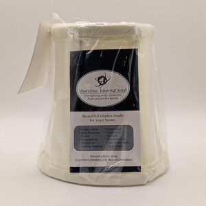 May include: A cream-colored lampshade in plastic wrap. The label reads "Shoreline International" and "Beautiful shades made for your home." Features include luxury fabric and rust-resistant frame.
