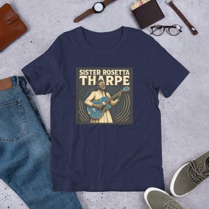 Puede incluir: Camiseta azul marino con un gráfico de Sister Rosetta Tharpe tocando una guitarra azul. El gráfico está en un cuadrado con el texto "SISTER ROSETTA THARPE" encima de la imagen. La camiseta se muestra sobre una superficie plana con vaqueros y accesorios.