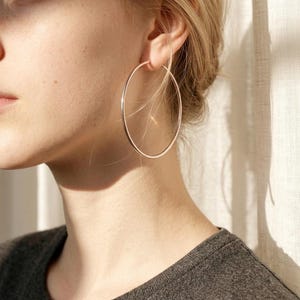 May include: Rose gold hoop earrings. The large, circular earrings are worn on the ear. The earrings are a simple, minimalist design. The model is wearing a grey top.