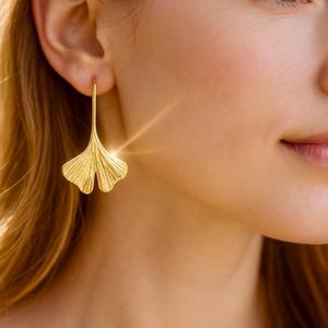 Op de afbeelding: Een goudkleurige oorbel in de vorm van een ginkgoblad. De oorbel hangt aan een dunne haak en heeft gedetailleerde nerven. Gedragen door een persoon met een lichte huid, met een vage achtergrond. Botanisch thema.