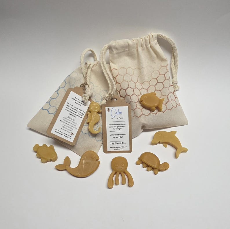 May include: A collection of natural beeswax sensory toys in the shape of sea creatures, including a fish, seahorse, dolphin, octopus, and turtle. The toys are light brown and are displayed with two drawstring bags with a honeycomb pattern.