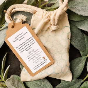 May include: A small beige drawstring bag with a tag attached. The tag reads "Smooth. Warm. Naturally honey-scented. Non-toxic." and lists uses for the tactile sensory fidget. The bag is on a bed of green leaves.