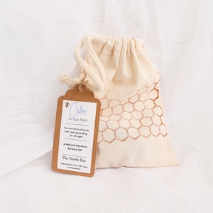 May include: A natural beeswax sensory set in a beige drawstring bag with a honeycomb pattern. A brown tag reads "Calm In Your Palm" and "The North Bee". The set is designed for focus and grounding.