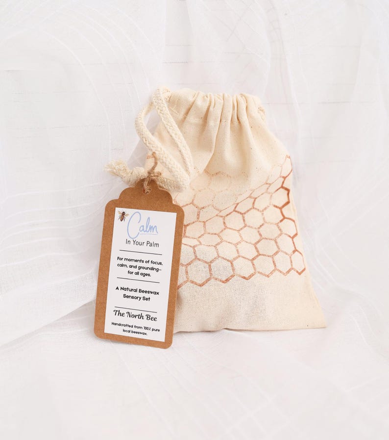 May include: A natural beeswax sensory set in a beige drawstring bag with a honeycomb pattern. A brown tag reads "Calm In Your Palm" and "The North Bee". The set is designed for focus and grounding.