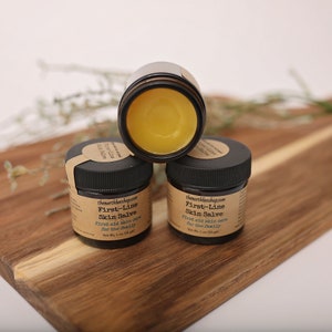 May include: Three small amber glass jars with black lids containing a yellow salve. The jars are labeled "First-Line Skin Salve" and "First aid skin care for the family".