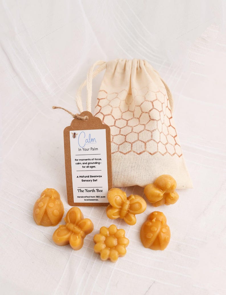 May include: A natural beeswax sensory set featuring various shapes, including a ladybug, butterfly, and flower. The set includes a drawstring bag with a honeycomb pattern and a tag that reads "Calm In Your Palm".