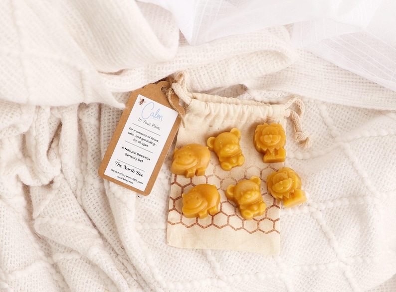May include: A natural beeswax sensory set with animal shapes, including bears and lions, in a small drawstring bag. The set is labeled "Calm in Your Palm" and is handmade from 100% pure food-grade beeswax.