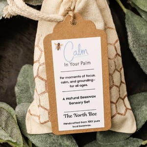 May include: A beige drawstring bag with a brown tag that reads "Calm In Your Palm." The tag also says "A Natural Beeswax Sensory Set" and "The North Bee." The bag is surrounded by green leaves.
