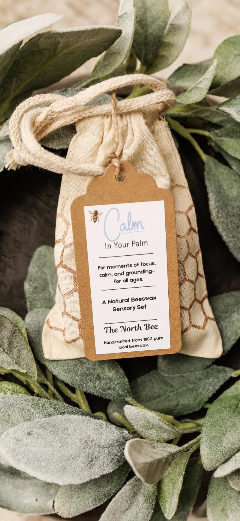 May include: A beige drawstring bag with a brown tag that reads "Calm In Your Palm." The tag also says "A Natural Beeswax Sensory Set" and "The North Bee." The bag is surrounded by green leaves.