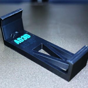 May include: A black plastic phone stand with the text "AD3D" printed on it. The stand is designed to hold a phone upright.