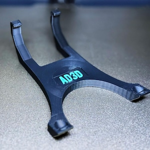 May include: A black plastic stand with a U-shaped design. The stand has the text "AD3D" printed on it in teal.