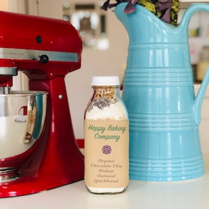 May include: A glass jar filled with dry ingredients for making organic chocolate chip walnut oatmeal quickbread. The jar has a brown paper label that reads "Happy Baking Company" and lists the ingredients. The jar is sitting on a counter next to a red stand mixer and a blue pitcher.