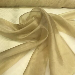 May include: A close-up of a flowing, sheer fabric in a light champagne color. The fabric is draped and folded, creating soft shadows and highlights. The material appears to be lightweight and suitable for various craft projects or apparel.