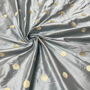 May include: A close-up of a gray fabric with a lustrous sheen. The fabric is adorned with large, evenly spaced, gold-colored polka dots. The material is gathered and folded, creating a swirling pattern.