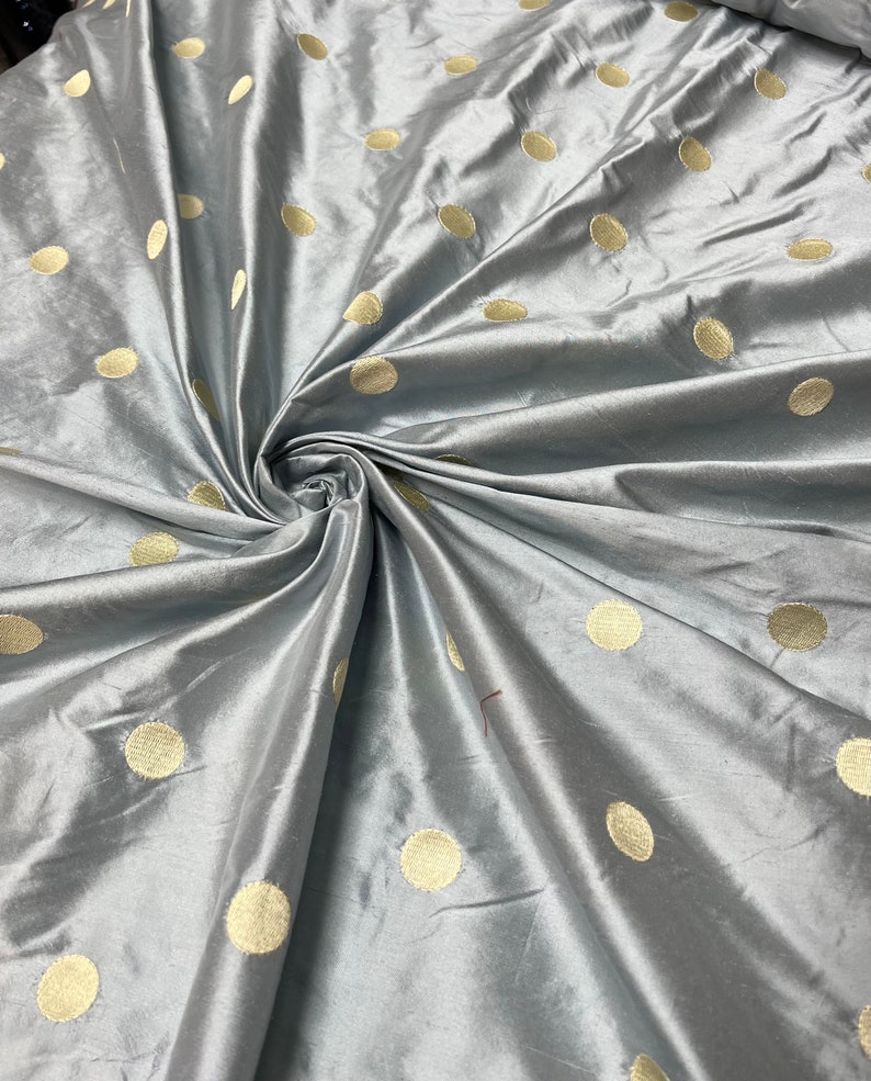 May include: A close-up of a gray fabric with a lustrous sheen. The fabric is adorned with large, evenly spaced, gold-colored polka dots. The material is gathered and folded, creating a swirling pattern.