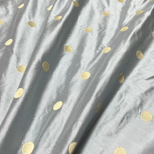 May include: A close-up of a light blue fabric with a pattern of gold-colored polka dots. The fabric has a subtle sheen and is slightly wrinkled, creating shadows and highlights. The polka dots are evenly spaced across the surface.