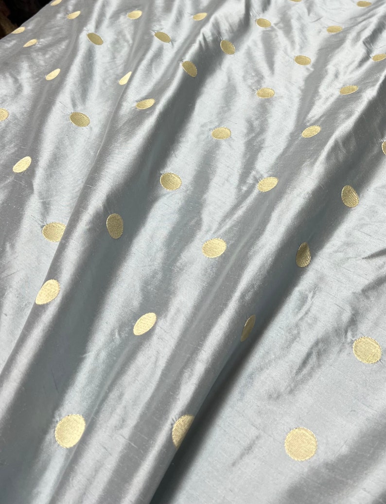 May include: A close-up of a light blue fabric with a pattern of gold-colored polka dots. The fabric has a subtle sheen and is slightly wrinkled, creating shadows and highlights. The polka dots are evenly spaced across the surface.