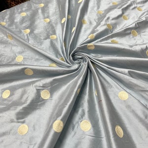 May include: A swatch of light blue fabric with a lustrous sheen, featuring a pattern of large, evenly spaced, golden-yellow polka dots. The fabric is gathered and swirled, showcasing its flowing texture and color variations.