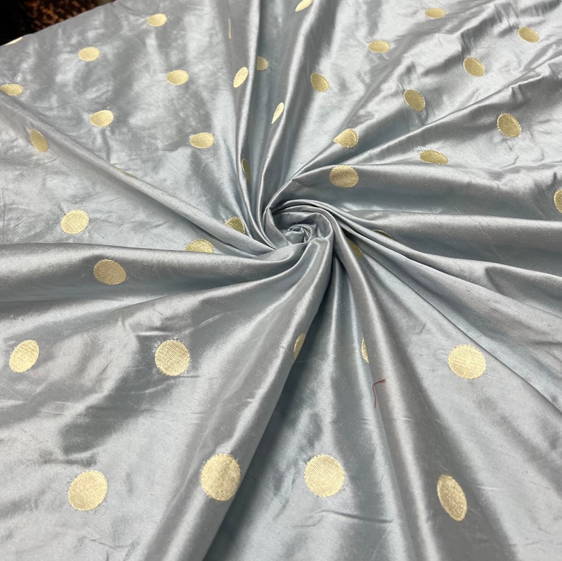 May include: A swatch of light blue fabric with a lustrous sheen, featuring a pattern of large, evenly spaced, golden-yellow polka dots. The fabric is gathered and swirled, showcasing its flowing texture and color variations.