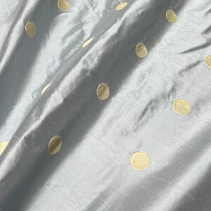 May include: Close-up of a light blue fabric with a satin sheen. The fabric is adorned with evenly spaced, large, gold-colored polka dots. The material is draped, creating soft folds and shadows, highlighting the texture and pattern.