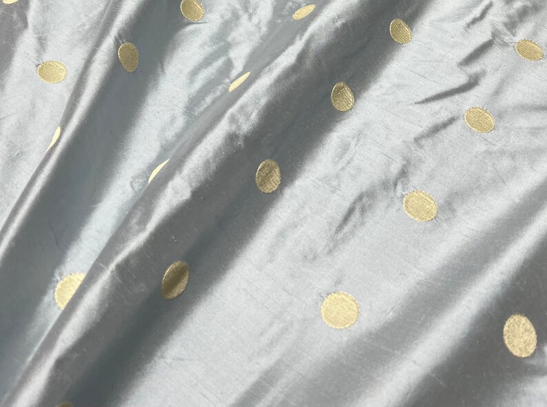 May include: Close-up of a light blue fabric with a satin sheen. The fabric is adorned with evenly spaced, large, gold-colored polka dots. The material is draped, creating soft folds and shadows, highlighting the texture and pattern.