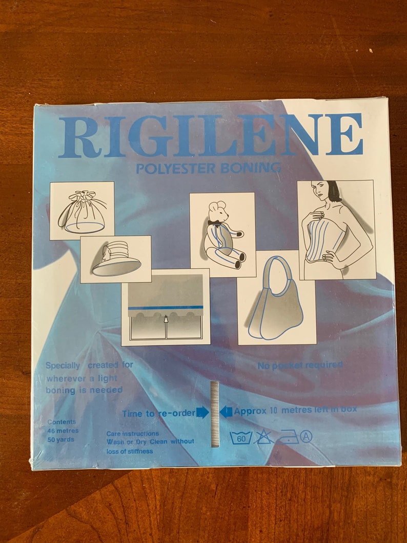 K&ouml;nnte beinhalten: Eine blaue Box mit dem Text "RIGILENE POLYESTER BONING" auf der Vorderseite. Die Box zeigt verschiedene M&ouml;glichkeiten, die Bonierung zu verwenden, darunter einen Hut, einen Teddyb&auml;r, eine Tasche und ein Korsett. Die Box enth&auml;lt auch Pflegehinweise und Informationen &uuml;ber den Inhalt, der 46 Meter oder 50 Yards Bonierung umfasst.