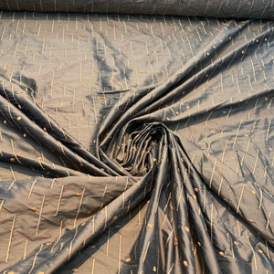May include: A dark grey fabric with a sheen, patterned with vertical lines and scattered dots in a lighter colour. The fabric is draped, showing folds and texture, suggesting it's a decorative textile, possibly for upholstery.