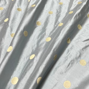 May include: A close-up of a light blue fabric with a subtle sheen. The fabric is adorned with evenly spaced, large, golden-yellow polka dots. The material is draped, creating soft folds and shadows, highlighting its texture and design.