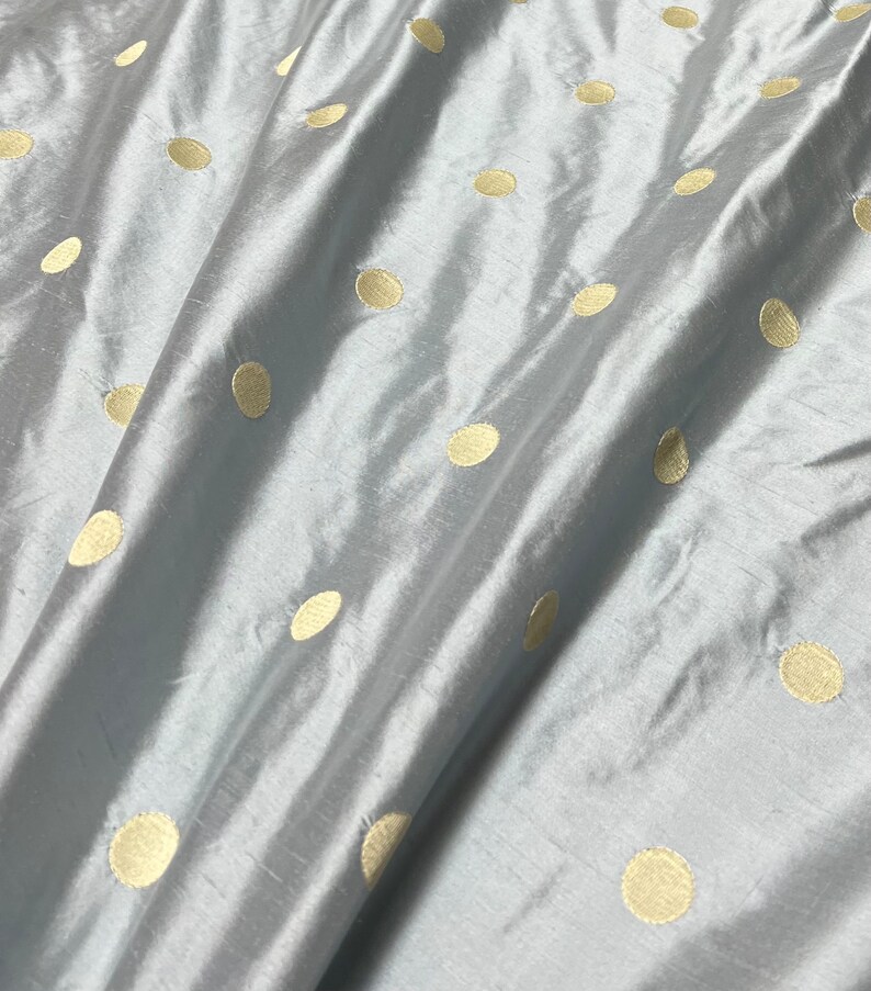 May include: A close-up of a light blue fabric with a subtle sheen. The fabric is adorned with evenly spaced, large, golden-yellow polka dots. The material is draped, creating soft folds and shadows, highlighting its texture and design.