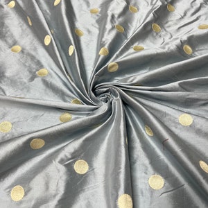 May include: Silky gray fabric with a pattern of large, evenly spaced, gold-colored polka dots. The fabric is folded and draped, creating shadows and highlights that emphasize its texture and sheen. Suitable for apparel or home decor.