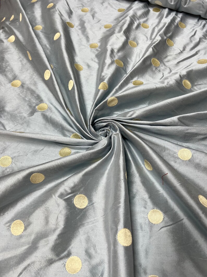May include: Silky gray fabric with a pattern of large, evenly spaced, gold-colored polka dots. The fabric is folded and draped, creating shadows and highlights that emphasize its texture and sheen. Suitable for apparel or home decor.
