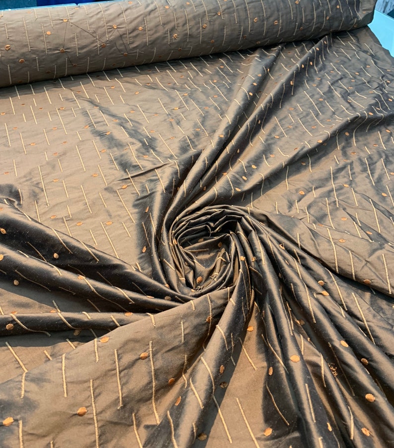 May include: A roll of brown fabric with a slight sheen. The material displays a pattern of vertical and diagonal lines, alongside small, evenly spaced dots in a lighter brown hue. The fabric is gathered in a swirl, highlighting its texture.