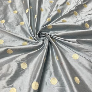 May include: A close-up shot of a gray fabric with a satin sheen. The fabric is adorned with large, evenly spaced, gold-colored polka dots. The fabric is gathered and folded, creating a textured appearance.