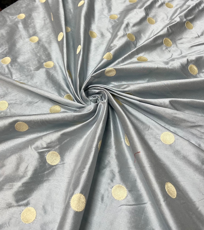 May include: A close-up shot of a gray fabric with a satin sheen. The fabric is adorned with large, evenly spaced, gold-colored polka dots. The fabric is gathered and folded, creating a textured appearance.