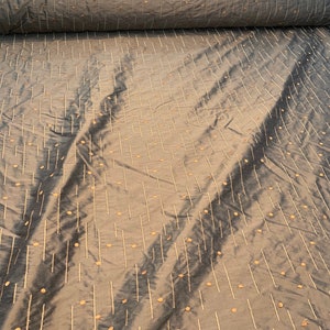 May include: A roll of grey-green fabric with a subtle sheen. The material is patterned with vertical lines and small, evenly spaced dots in a lighter beige hue. The fabric has a soft, flowing appearance, suggesting it is lightweight and drapable.