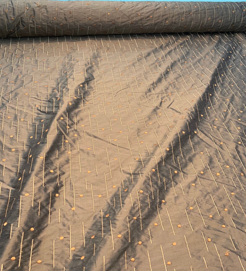 May include: A roll of grey-green fabric with a subtle sheen. The material is patterned with vertical lines and small, evenly spaced dots in a lighter beige hue. The fabric has a soft, flowing appearance, suggesting it is lightweight and drapable.