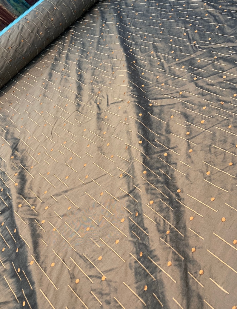May include: A roll of grey fabric with a subtle sheen. The fabric is decorated with a pattern of diagonal lines and small, evenly spaced dots in a lighter, contrasting colour. The fabric appears to be soft and pliable.