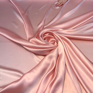 May include: A close-up of a soft, smooth, blush pink satin fabric. The fabric is draped and folded, creating a swirl pattern.