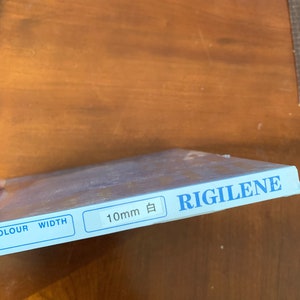 K&ouml;nnte beinhalten: Eine blaue und wei&szlig;e Schachtel mit dem Text "RIGILENE" und "10mm 白" darauf gedruckt. Die Schachtel hat auch die W&ouml;rter "COLOUR" und "WIDTH" aufgedruckt.